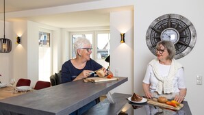 Un comedor moderno con una mesa larga, dos personas comiendo y un reloj de pared.