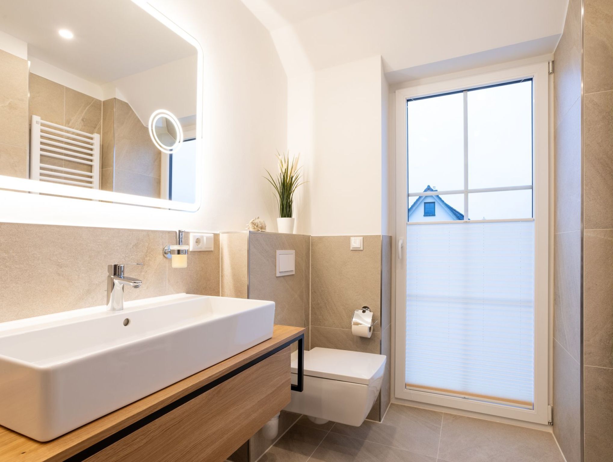 A modern bathroom with a large white sink, a mirror with built-in lights, a toilet, and a window with blinds.