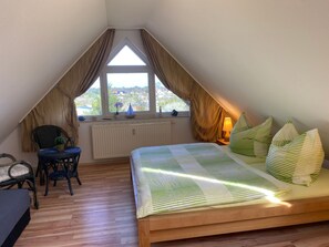 Un dormitorio con techo inclinado, una cama con ropa de cama a rayas, una silla, un escritorio y una ventana con cortinas.