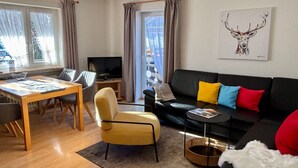 A modern living room with a wooden dining table, a yellow armchair, a black sofa with colorful cushions, a television on a stand, and a large window with curtains. - Haus Rosenwirth by Interhome (Mittenwald)