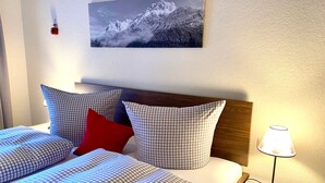 A hotel room with a bed, checkered bedding, a red pillow, a nightstand with a lamp, and a framed picture of a mountain on the wall. - Haus Rosenwirth by Interhome (Mittenwald)
