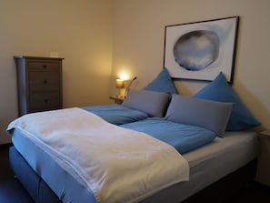 Ein ordentlich bezogenes Bett mit blau-weißer Bettwäsche, ein Nachttisch mit Lampe und ein an der Wand hängendes Bild.