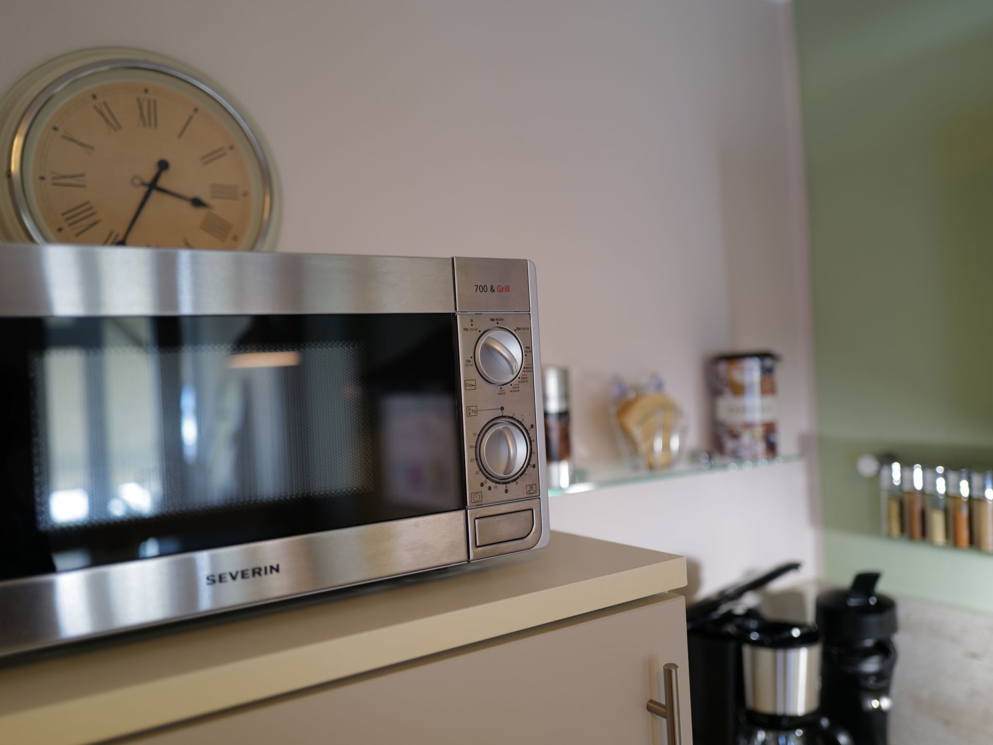 Une cuisine avec un four à micro-ondes posé sur un meuble, une horloge au mur et une étagère remplie d’ustensiles de cuisine.