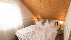 Une chambre avec un plafond en bois incliné, un lit recouvert de draps blancs, deux lampes de chevet et une fenêtre ornée de rideaux légers.