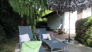 Zwei Liegestühle mit Kissen, eine grüne Decke und ein Sonnenschirm in einem Garten mit Bäumen und einem Gebäude im Hintergrund.