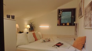 Ein kleines, gemütliches Schlafzimmer mit einem Einzelbett, einem Nachttisch mit Lampe, einem Fenster mit Ausblick und einer Wandablage mit Dekoration.