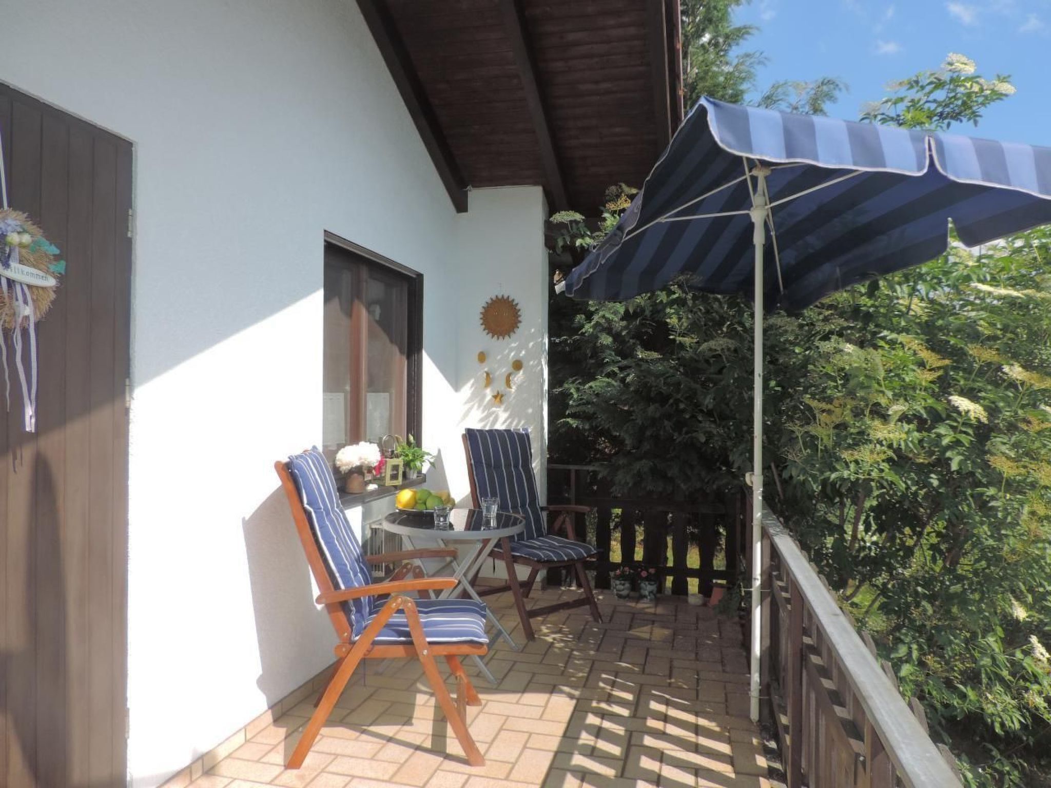 Un balcone con due poltrone a righe, un tavolino con un vaso e della frutta, e un ombrellone a righe blu.