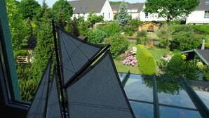 Vista pela janela, com uma estrutura triangular semelhante a uma vela em primeiro plano, um jardim com diversas plantas e flores, e casas ao fundo.