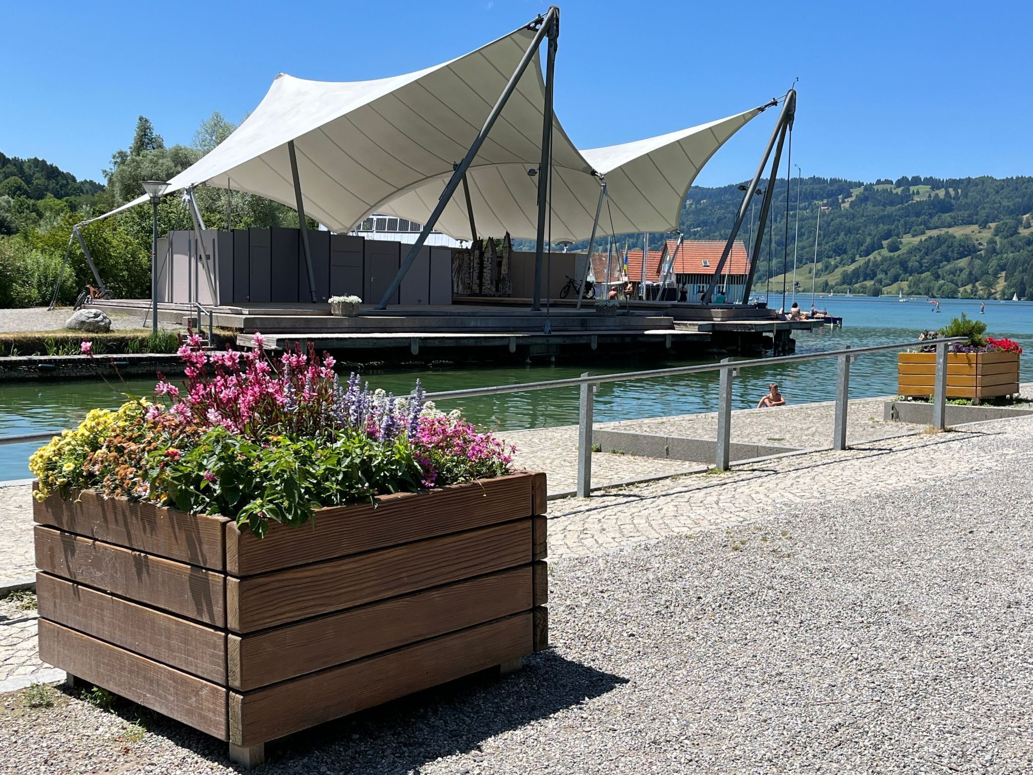 A waterfront area with a wooden planter of flowers, a covered seating area, and a view of the lake.
