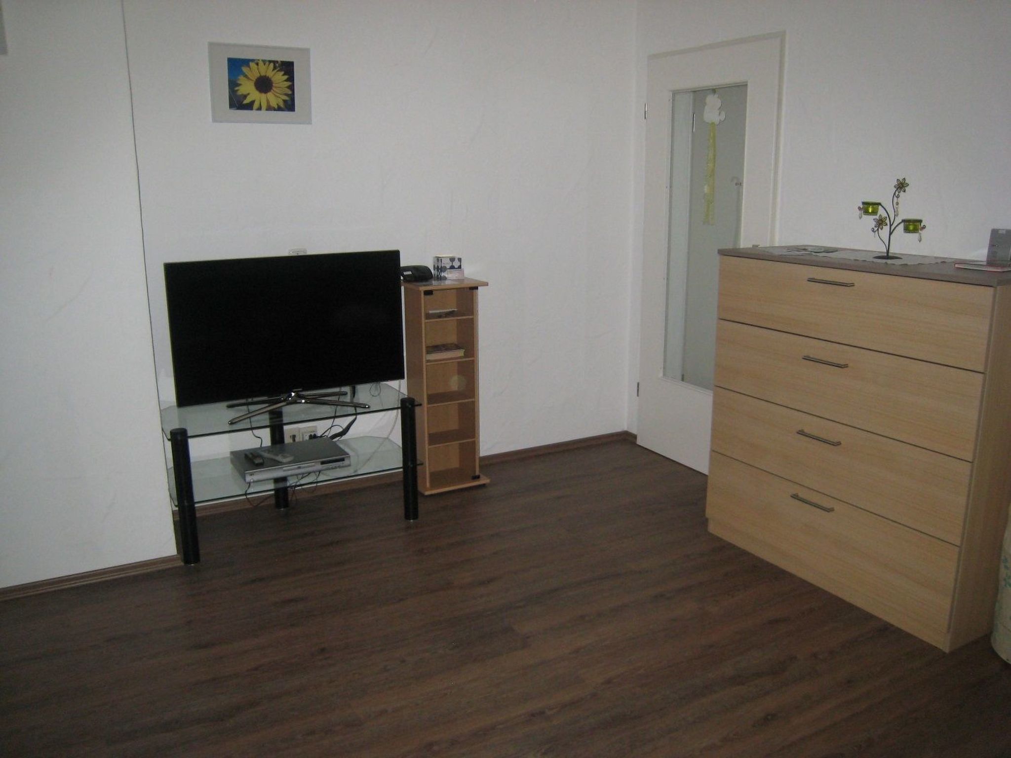 Una habitación con una televisión de pantalla plana sobre un pedestal de vidrio, un mueble de madera con cajones y una puerta con paneles de vidrio esmerilado.