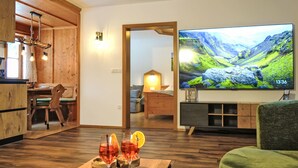 Een gezellige woonkamer met houten vloer, een televisie waarop een panoramisch landschap te zien is, en een eethoek met houten meubels.