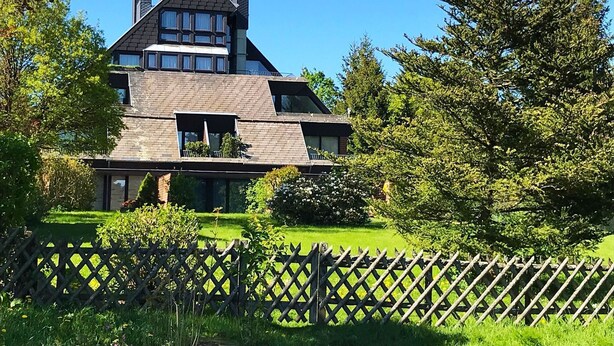 Une maison moderne à deux étages, dotée d’un toit à forte pente, de grandes fenêtres et d’une cour clôturée.