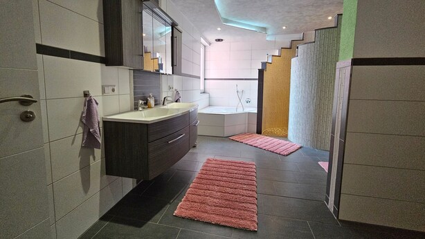 Ein modernes Badezimmer mit einem großen Waschtisch, einem Spiegel, einer Dusche und einer Treppe, die in einen anderen Bereich führt.
