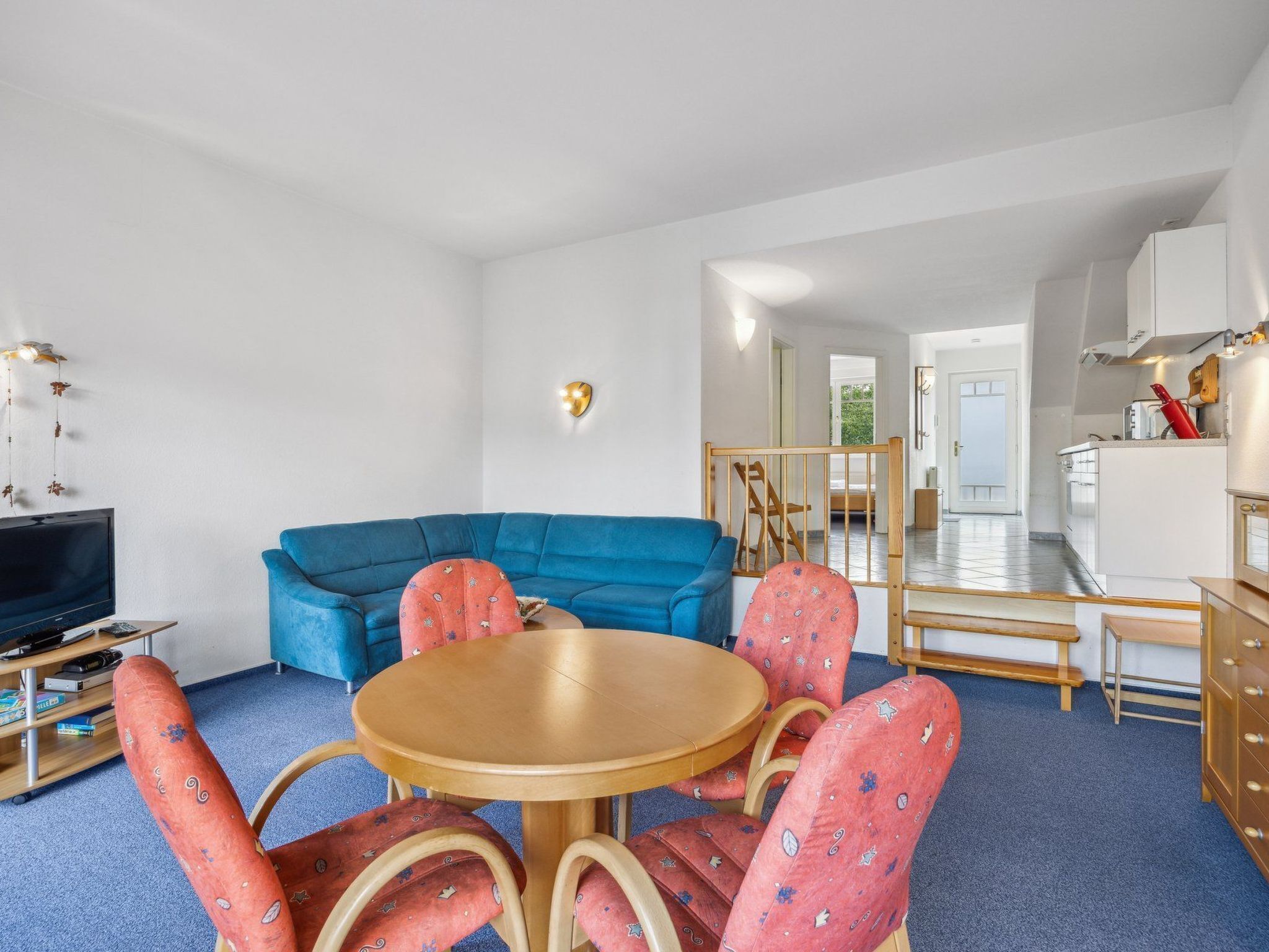 Un espacio habitable pequeño con un sofá azul, una mesa redonda de madera con sillas a juego, un televisor y una kitchenette.