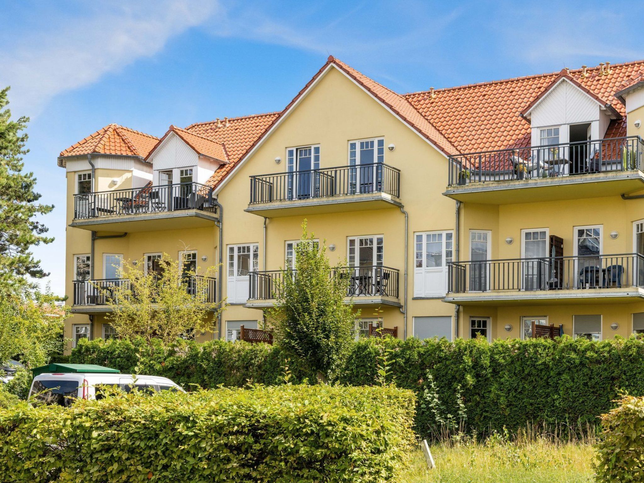 A multi-story residential building with balconies, a car parked in front, and well-maintained hedges.