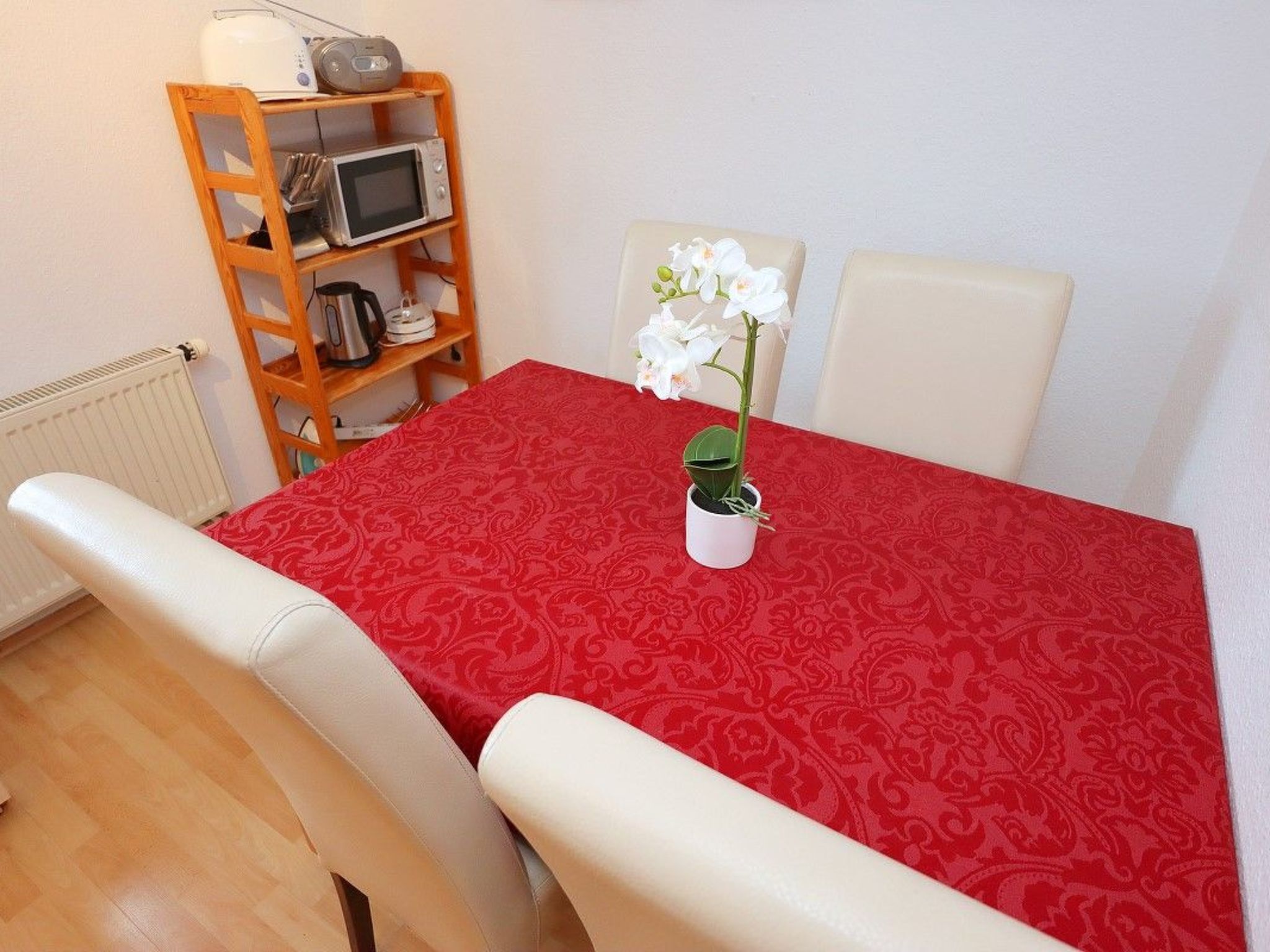 A dining area with a red patterned tablecloth, a white chair, a small potted orchid, and a wooden shelf with kitchen appliances.