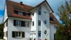 Una casa a due piani con tetto rosso, pareti bianche e un portico.