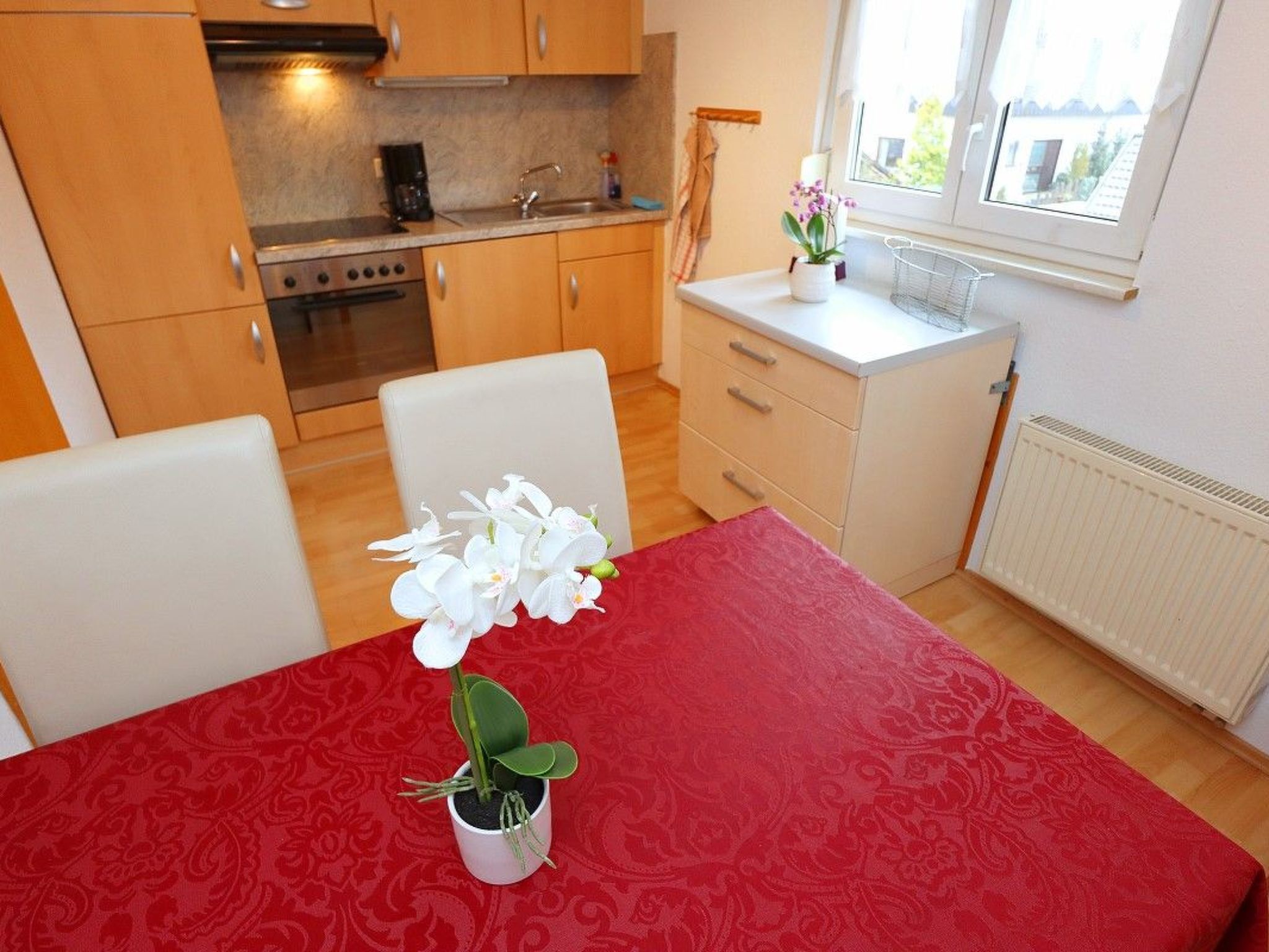 A compact kitchen with wooden cabinets, a stove, and a sink. There's a dining table with a red tablecloth and a potted orchid.