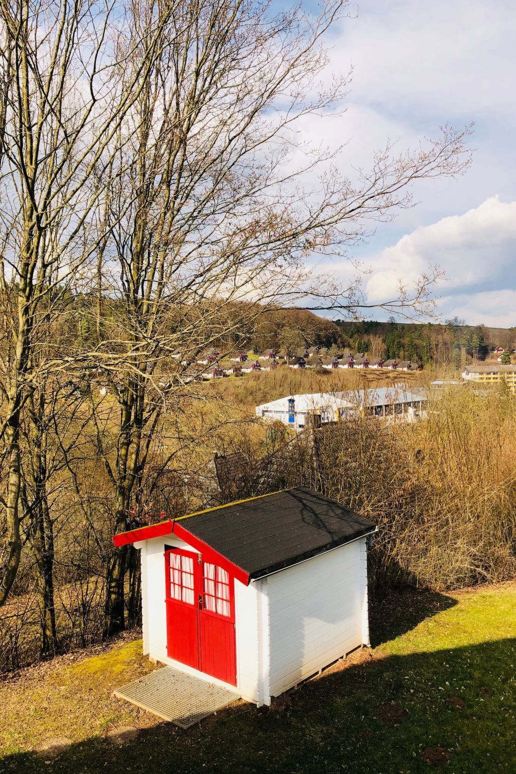 Ein kleines, weißes Gebäude mit roter Tür und schwarzem Dach, umgeben von Bäumen und einer grünen Wiese.