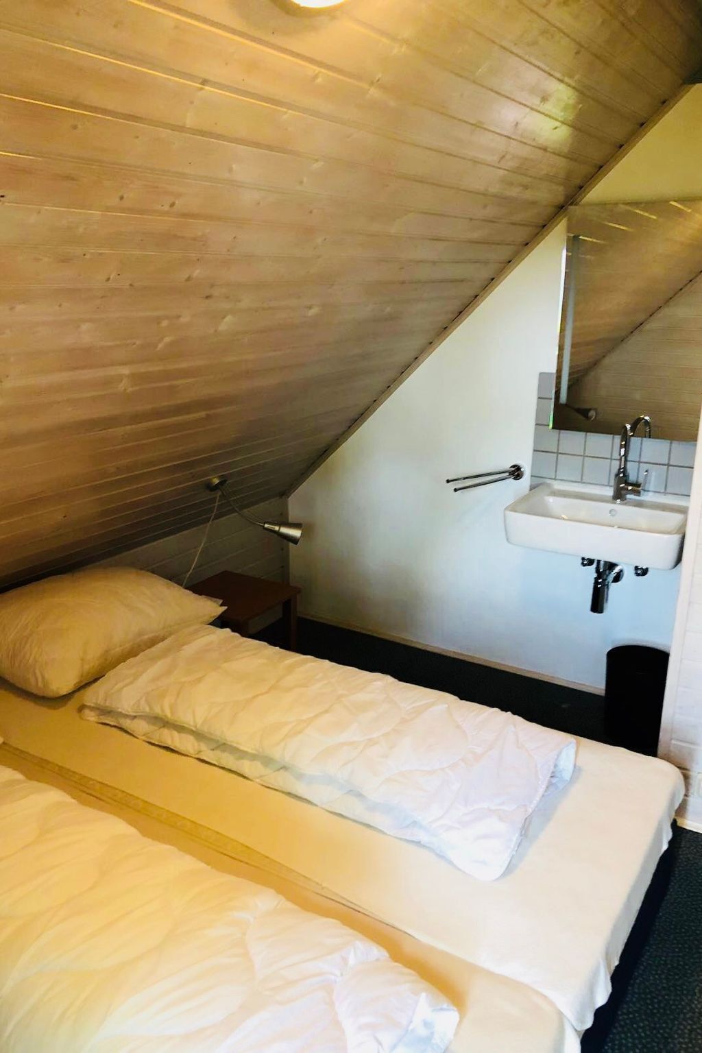 A small, single-bed room with a sloped ceiling, a wooden floor, a white sink, and a mirror above it.