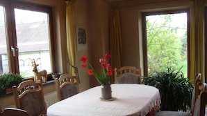 Un comedor con una mesa redonda cubierta con mantel blanco, rodeada de sillas de madera. Hay un jarrón con flores rojas sobre la mesa.
