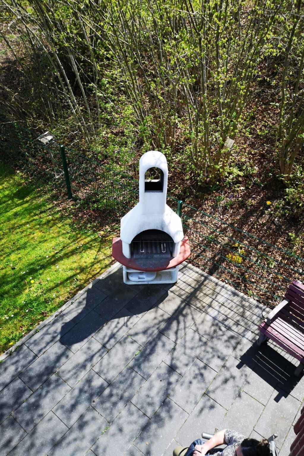 Una parrilla exterior blanca con chimenea, rodeada por un jardín con árboles y un área pavimentada.