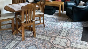 Una sala de estar con una mesa de comedor de madera, dos sillas, un sofá de cuero negro con cojines rojos y negros, y un suelo de baldosas estampadas.