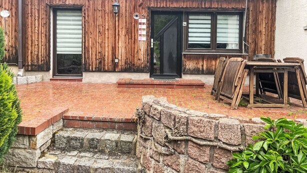 Un edificio rústico con fachada de madera, dos puertas, un pequeño espacio con mesas al aire libre y un muro de piedra.