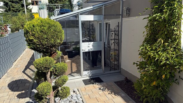 Een modern huis met een glazen deur die uitkomt op een terras, omringd door een tuin met diverse planten en een klein beeldje.
