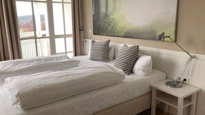 Un lit double bien fait, avec du linge de lit blanc et deux coussins à motifs, une table de chevet avec une lampe, et un grand tableau abstrait accroché au mur.