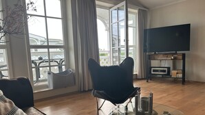 En stue med sovesofa, sort læderstol, fladskærms-tv og bogreol samt et sofabord i glas.