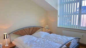 Un dormitorio con cama, dos lámparas de noche, una ventana con persianas y una mesita de noche con un objeto estampado con motivos florales.