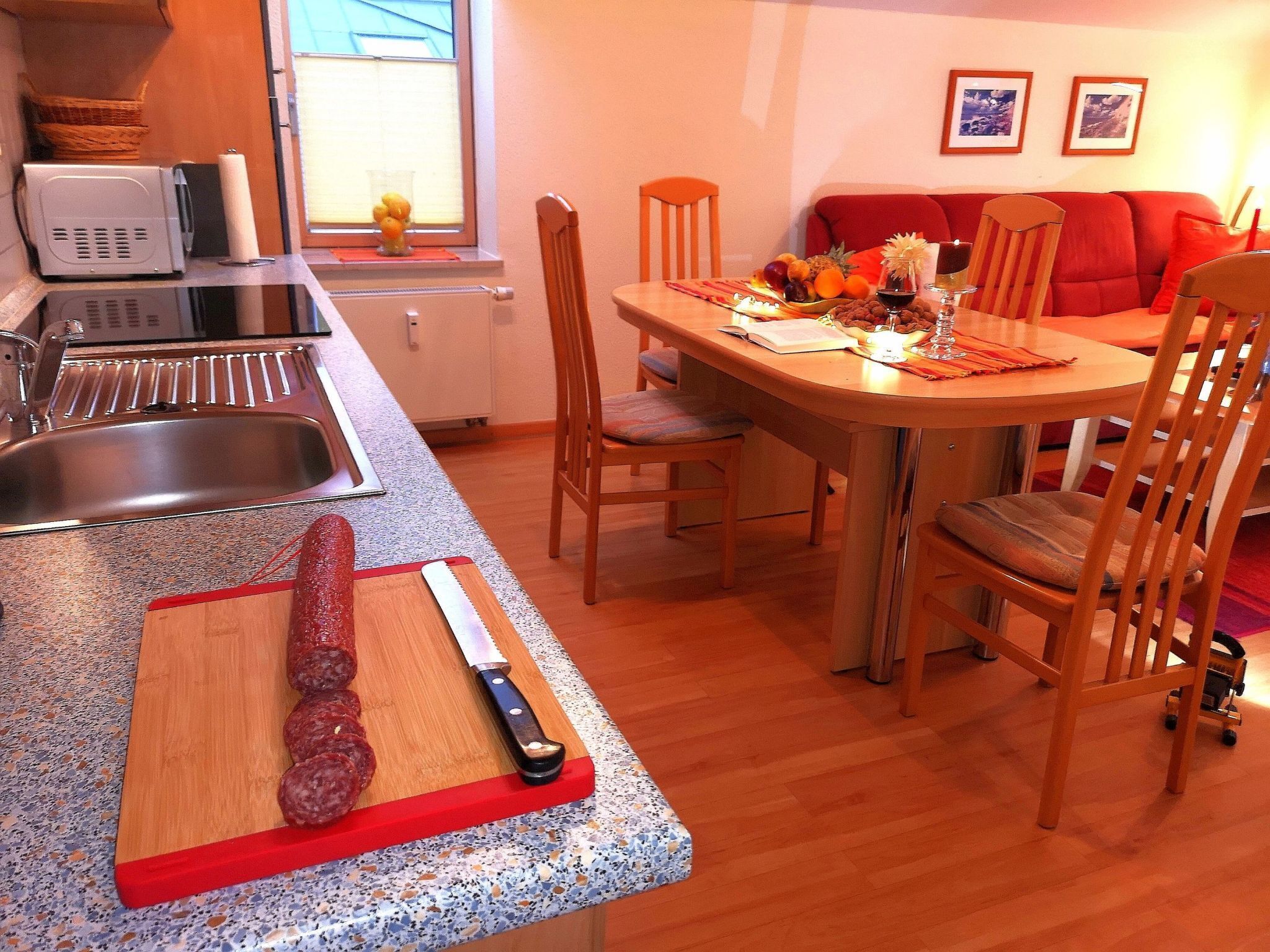 Una cocina con encimera de granito, tabla de madera con carne y un cuchillo. Se ve una mesa de comedor con sillas y un sofá rojo al fondo.