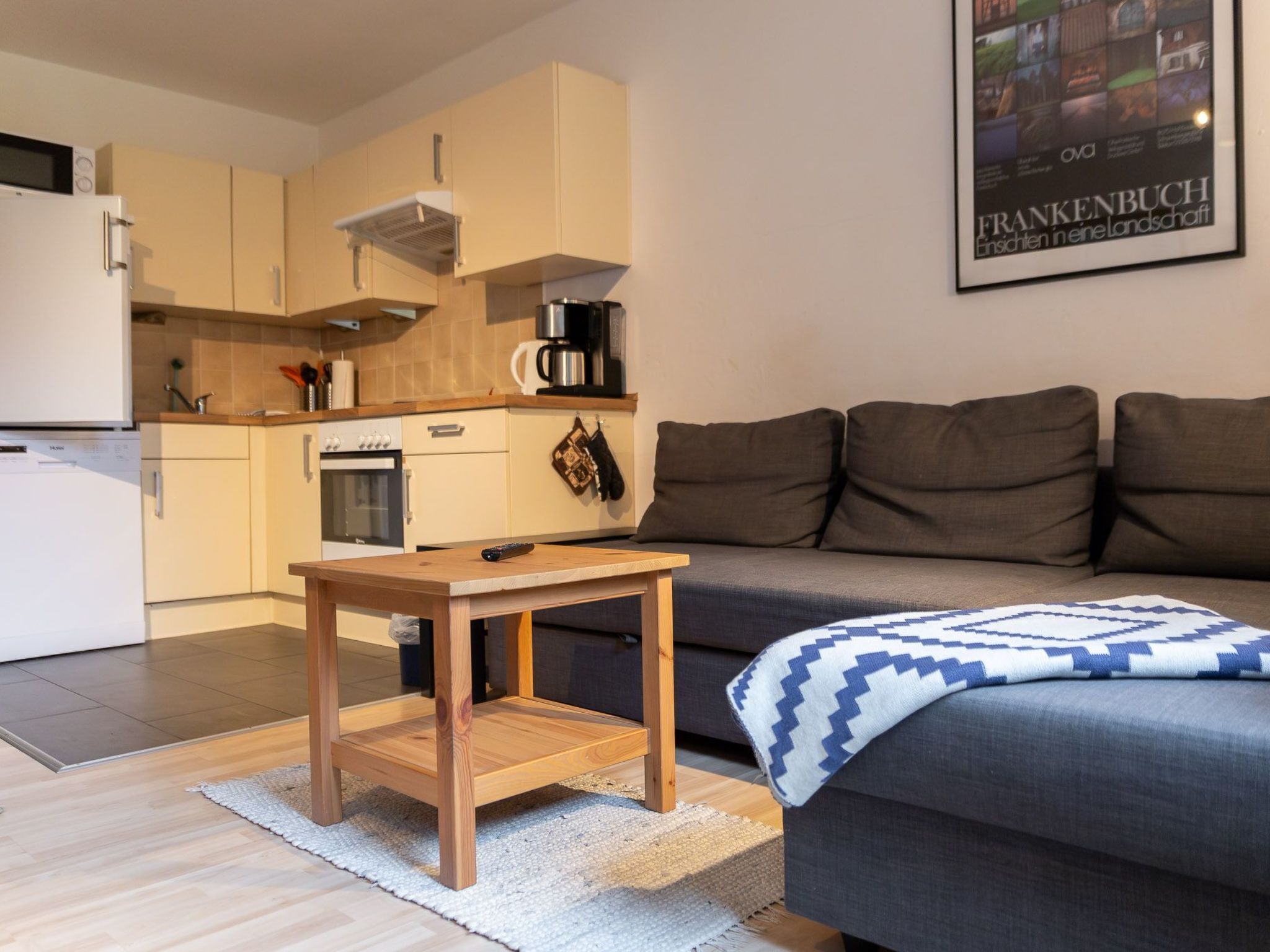 A modern kitchen with a built-in oven, a coffee machine, and a microwave. There is a wooden table and a grey sofa with a blue and white patterned throw blanket.