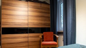 A bedroom with a wooden wardrobe, a single chair, a bed with patterned bedding, and a window with curtains. - mit zwei Schlafzimmern und Terrasse by Interhome (Weißenstadt)