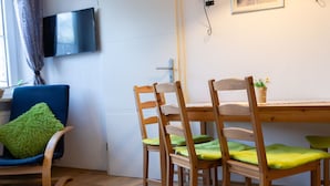 En matplats med ett träbord och stolar, en tv som är monterad på väggen och ett fönster med gardiner.