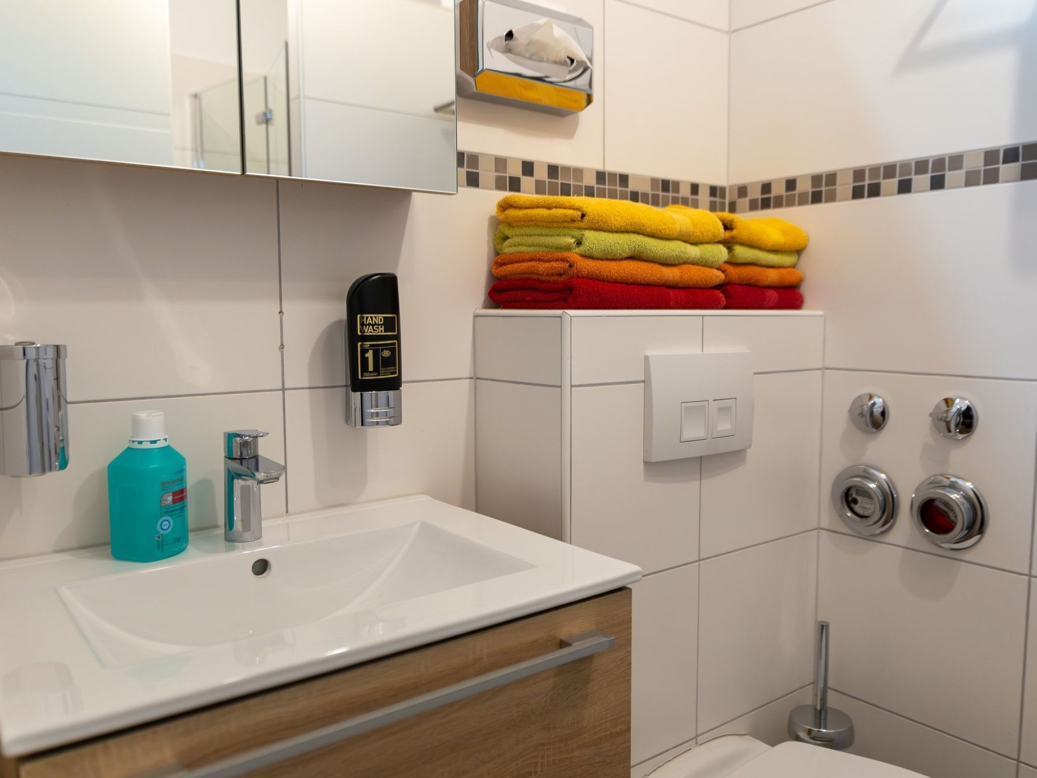 Un baño moderno con un lavamanos blanco, estación de lavado de manos, toallero con toallas de colores y un inodoro con bidé.