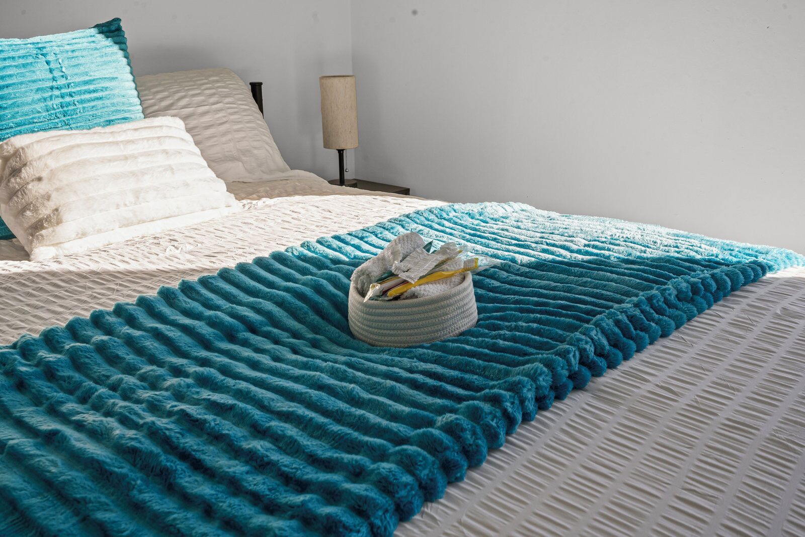 A neatly made bed with a blue and white striped blanket, a small round storage bin, and a bedside lamp on a white wall.