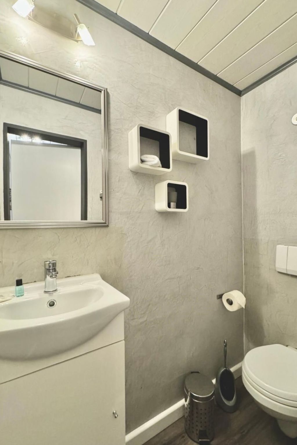 Un baño con lavamanos blanco, un espejo, tres repisas en la pared, inodoro y un cesto de basura.