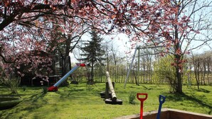 Ein Spielplatz mit Rutsche und Schaukeln, umgeben von Bäumen und einem Sandkasten mit Schaufeln und Eimern.