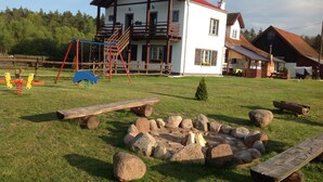 A rustic house with a red roof and white walls, a wooden deck, and a fire pit in the yard.