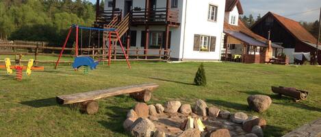 A rustic house with a red roof and white walls, a wooden deck, and a fire pit in the yard.