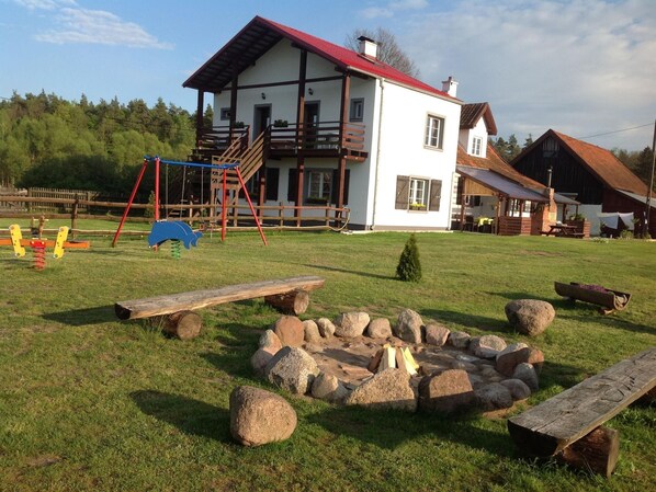 A rustic house with a red roof and white walls, a wooden deck, and a fire pit in the yard.