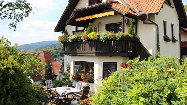 Een twee verdiepingen tellend huis met een balkon versierd met bloemen, een tuin met een tafel en stoelen, en uitzicht op de omgeving.