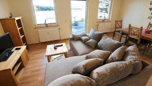 A living room with a sofa, a coffee table, a TV stand, and a dining table with chairs. There are three windows providing natural light. - Apartment Karpfen 4 by Interhome (Neukloster)