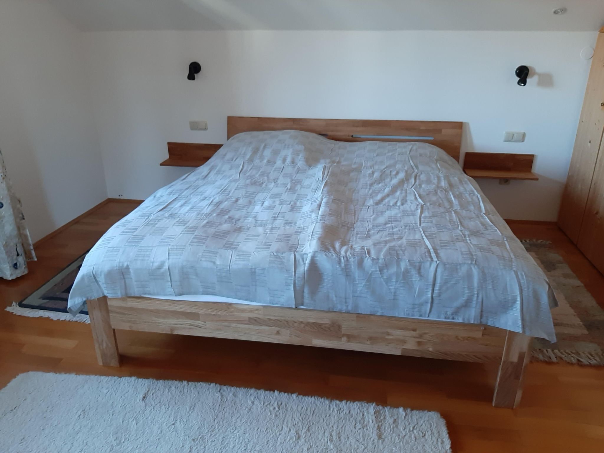 Ein Schlafzimmer mit einem hölzernen Bettgestell, einer weißen Bettdecke und einem kleinen Holzregal an der Wand.