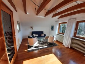 A spacious living room with a dark blue sofa, a coffee table, and two armchairs. There is a large window providing natural light and a view of trees outside. - Lechrauschen Landsberg by Interhome (Landsberg)