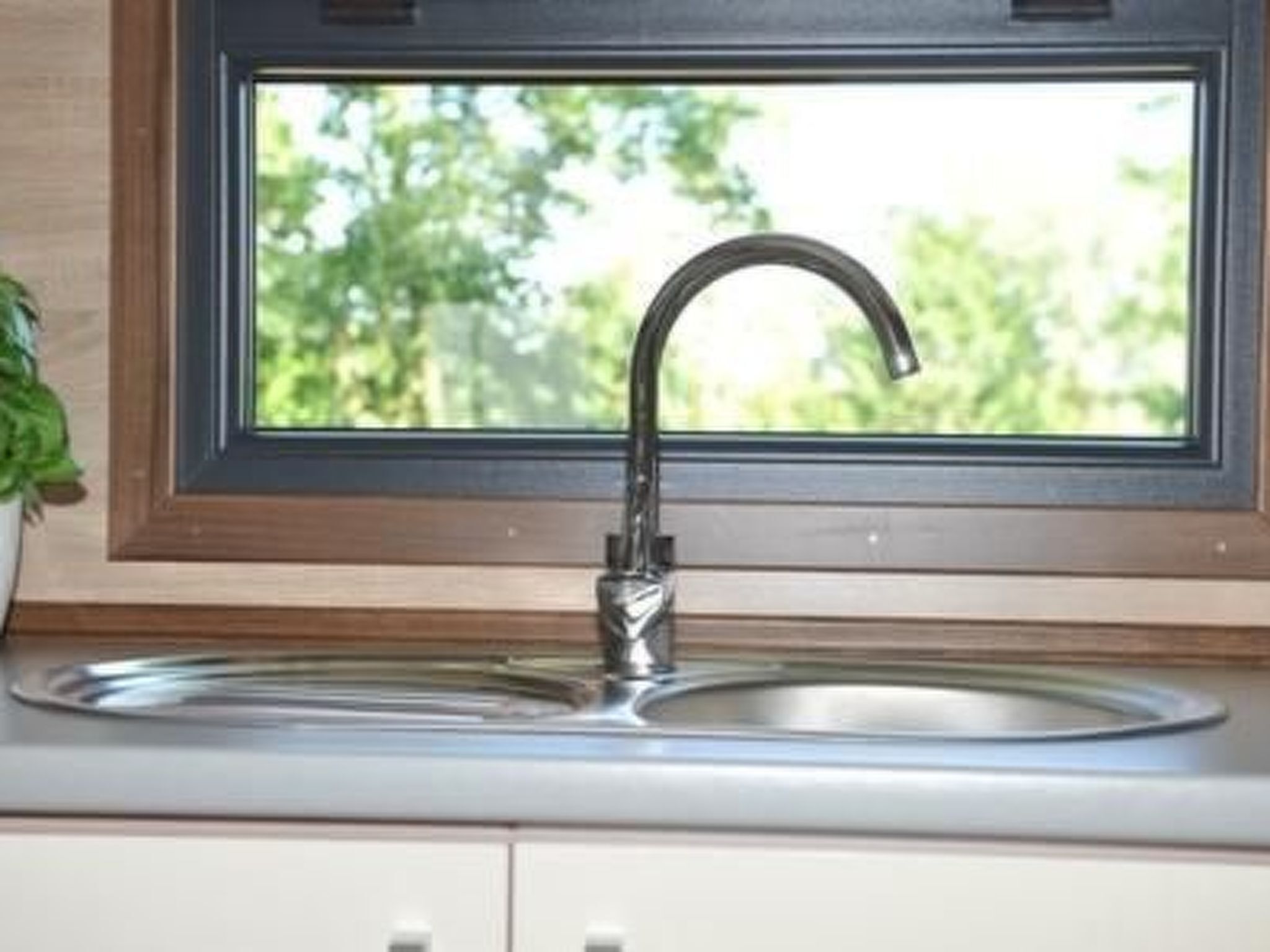 A kitchen with a stainless steel sink, a window above the sink, and a potted plant on the left.