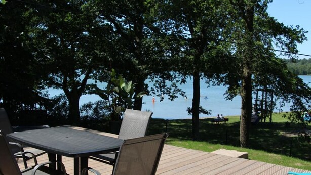 An outdoor deck with a table and chairs, overlooking a lake with trees and a grassy area. - Mobilheim Zweiundvierzig in Lychen by Interhome (Lychen)