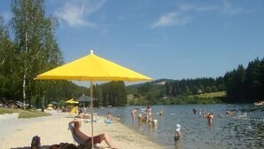 Une plage au bord d’un lac où l’on voit des gens nager et prendre des bains de soleil, un parasol jaune et un bateau sur l’eau.